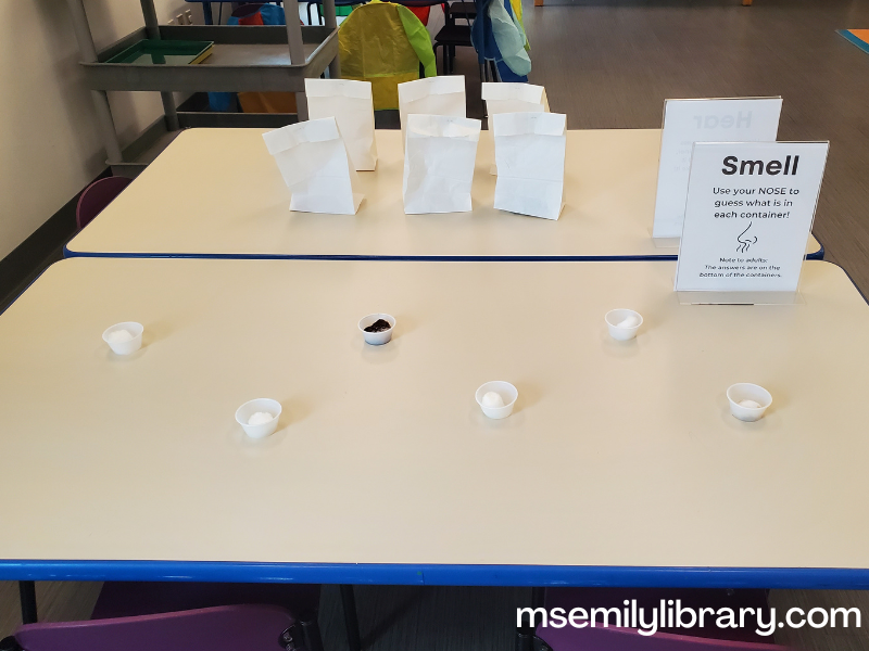 Smell station, showing six small condiment cups. Each has a cotton ball - the sign prompts participants to smell each and guess what it is, with answers written on the bottom of the cups.