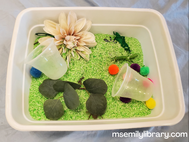 bug sensory bin showing green colored rice in a bin with fake flowers, foliage, pom poms, and plastic bugs.