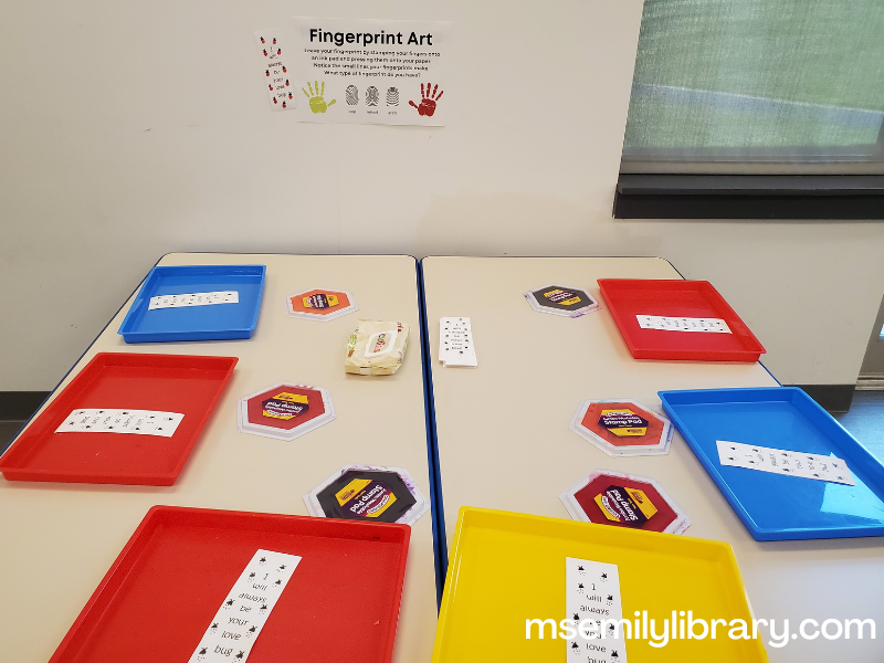 finger print art station, showing messy trays holding a bookmark with ladybug heads and spots that says "I will always be your love bug." Large stamp pads are on the table, as well as a package of wet wipes.