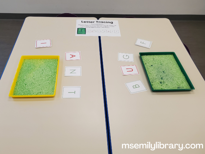 letter tracing, showing green colored rice in shallow messy trays. letter cards above the trays spell A N T and B U G, with other letter cards to the side.