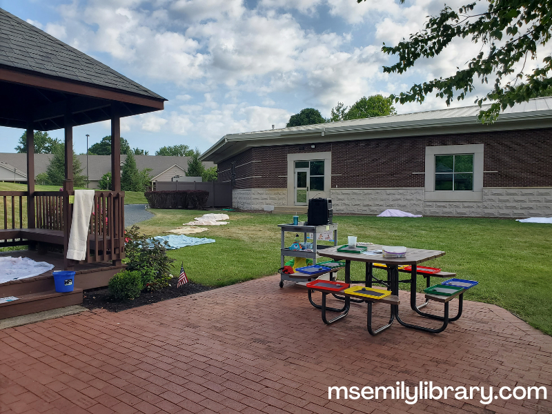 wide view of outdoor storytime area, showing gazebo to the left, picnic table to the right on bricked area, with a large grassy area beyond, with sheet-covered stations along the left and across the back near the library building in the background