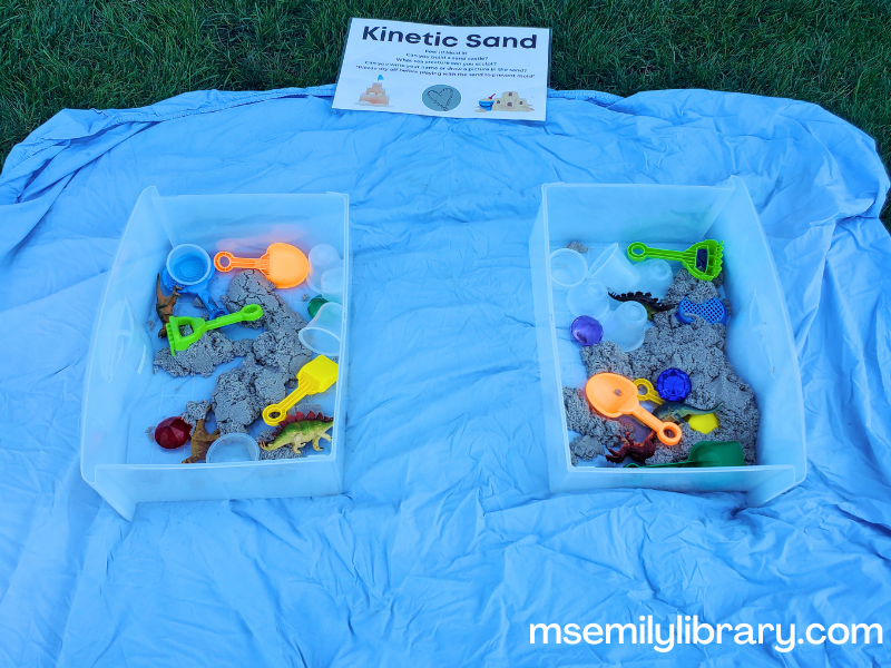 kinetic sand bins on a fitted sheet, with shovels, large plastic jewels, dinosaur toys, etc
