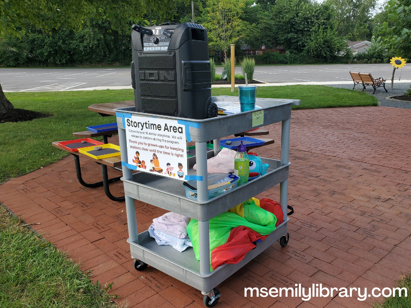 storytime cart, (large flat cart) with large parachute on the bottom, a basket of personal items such as sunscreen, bubble machine on the middle shelf, and a water cup and large personal PA speaker on the top.