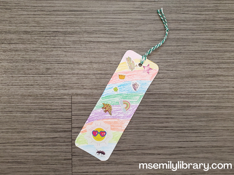 bookmark craft, showing a rainbow colored (with crayon) background with various stickers on a bookmark with a green tail.